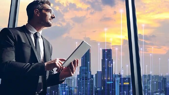 A man looking out of a skyscraper window holding a tab in his hand overlaid with graphics of transmission nodes
